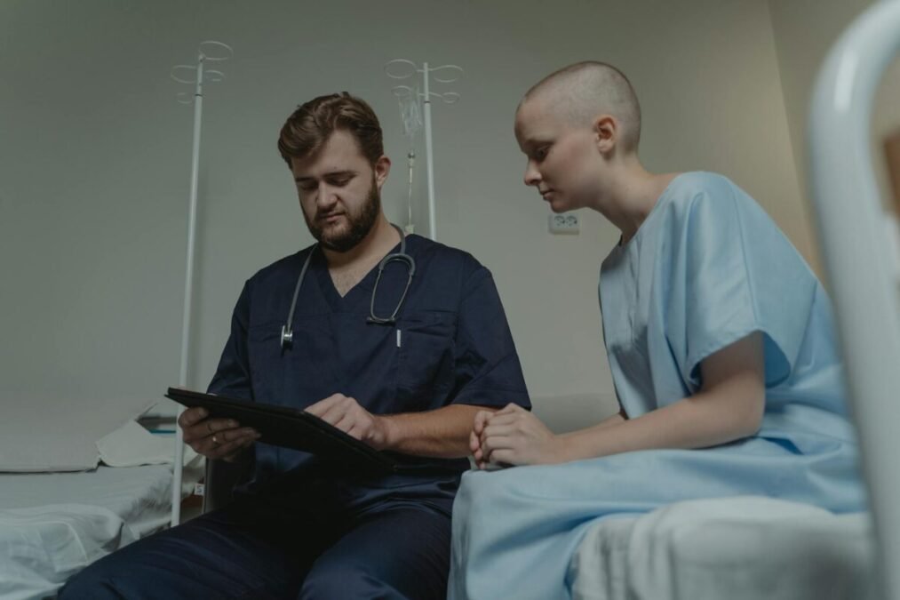 A doctor sits with a patient in a hospital room, reviewing information together on a tablet.